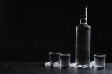 Photo of Bottle of vodka and shot glasses with ice on table against black background. Space for text