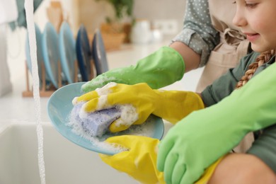 Photo of Mother and daughter wearing protective gloves washing plate above sink in kitchen, closeup