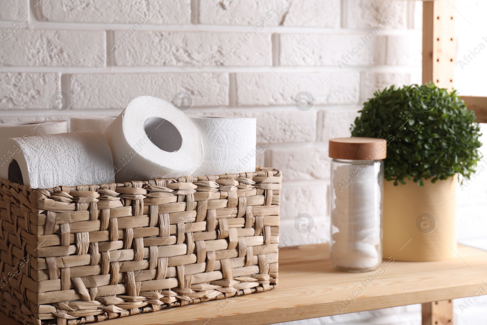 Photo of Toilet paper rolls in wicker basket, floral decor and cotton pads on wooden shelf near white brick wall