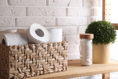 Photo of Toilet paper rolls in wicker basket, floral decor and cotton pads on wooden shelf near white brick wall