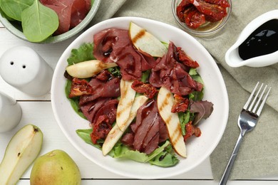 Photo of Delicious bresaola salad with sun-dried tomatoes, pear and balsamic vinegar served on white wooden table, flat lay