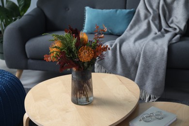 Photo of Vase with bouquet of beautiful flowers, book and glasses on wooden nesting tables near grey sofa indoors