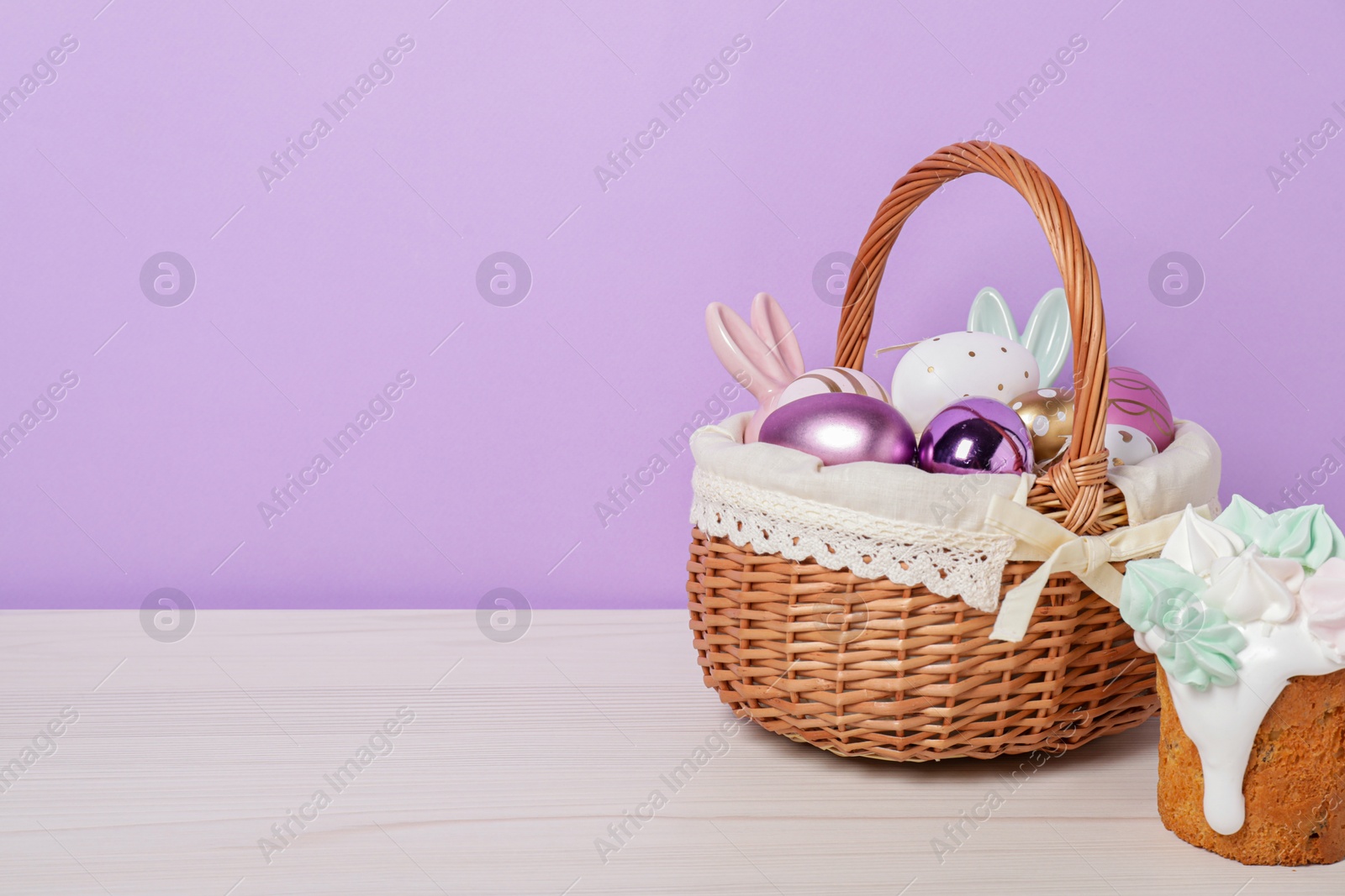 Photo of Easter basket with painted eggs, decorations and tasty cake on white wooden table. Space for text