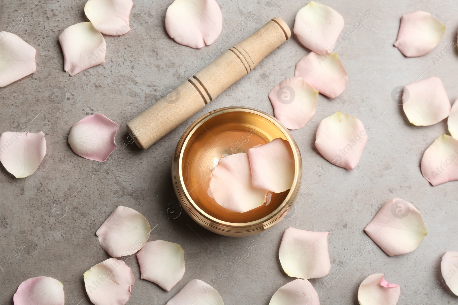 Photo of Golden singing bowl with petals and mallet on grey table, flat lay. Sound healing