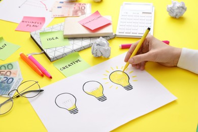 Photo of Woman drawing lamp on paper at yellow table, closeup. Startup concept