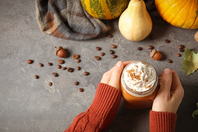 Woman holding mason jar of tasty pumpkin spice latte at grey table, top view