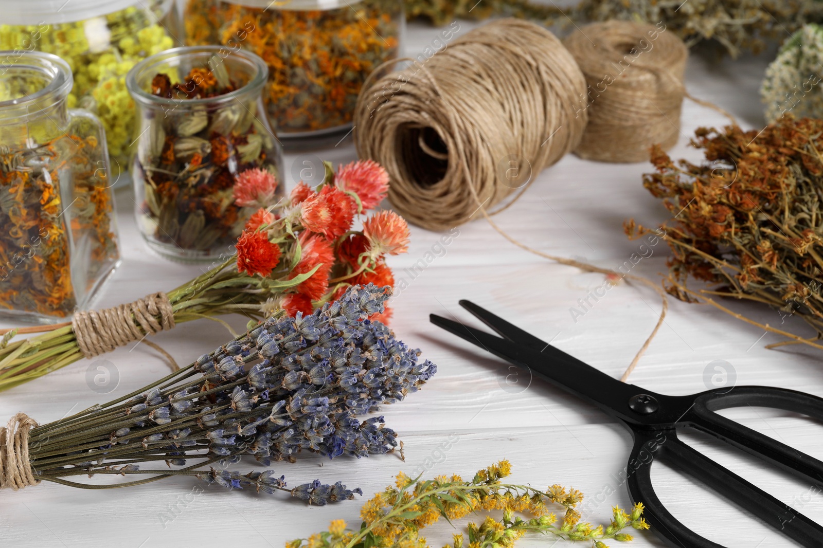 Photo of Bunches of dry flowers, different medicinal herbs, scissors and spools on white wooden table