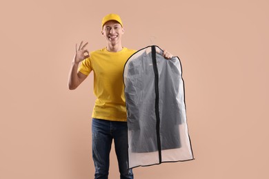 Dry-cleaning delivery. Happy courier holding garment cover with clothes and showing OK gesture on beige background