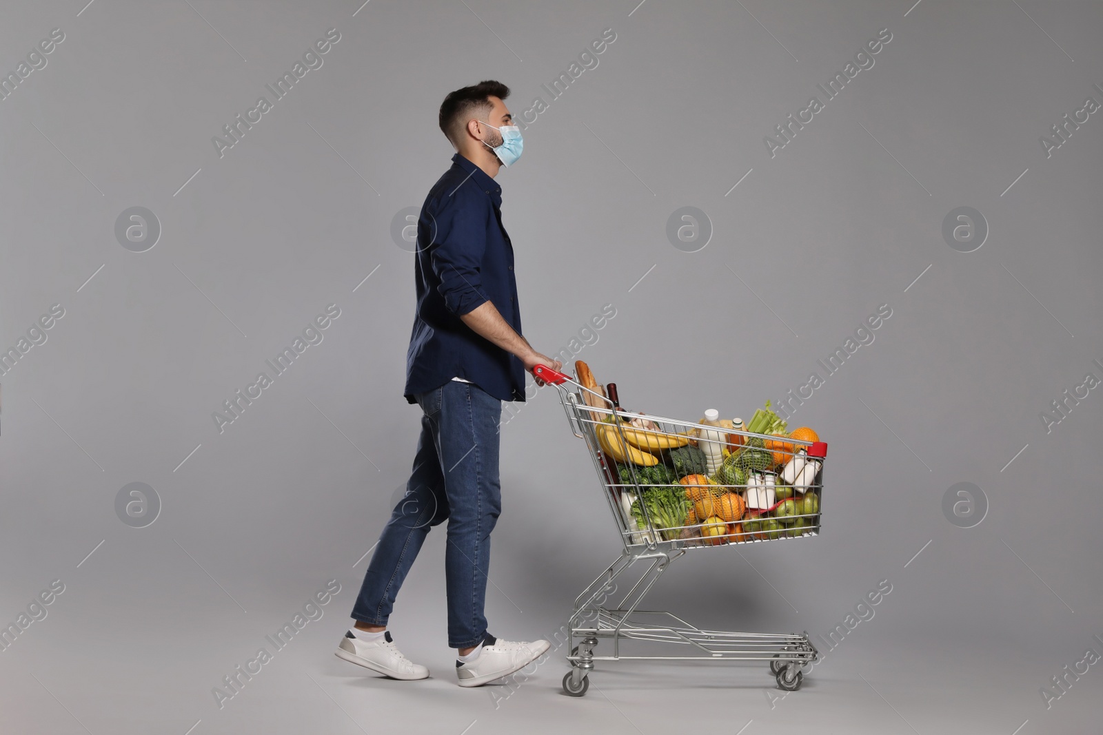 Photo of Man with protective mask and shopping cart full of groceries on light grey background