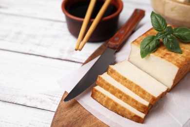 Photo of Board with smoked tofu, knife, basil and soy sauce on white wooden table. Space for text