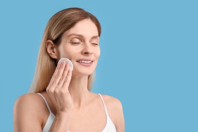 Beautiful woman removing makeup with cotton pad on light blue background, space for text