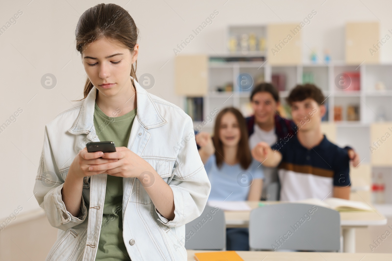 Photo of Teen problems. Lonely girl with smartphone standing separately from other students in classroom