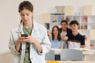 Teen problems. Lonely girl with smartphone standing separately from other students in classroom