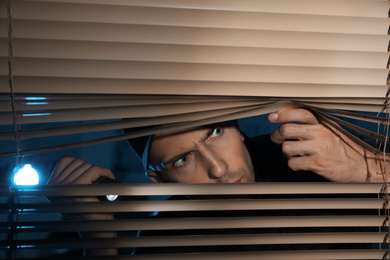 Male security guard with flashlight looking through window blinds in darkness