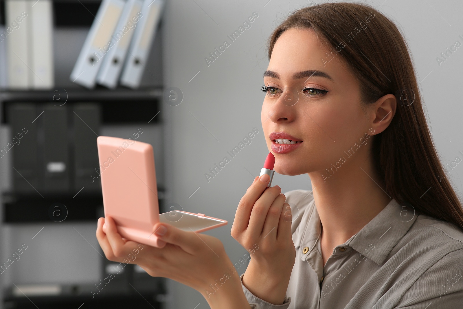 Photo of Young woman with cosmetic pocket mirror applying lipstick indoors