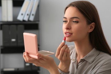 Young woman with cosmetic pocket mirror applying lipstick indoors