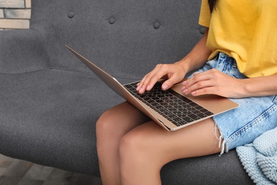 Photo of Young woman with modern laptop sitting on sofa at home, closeup