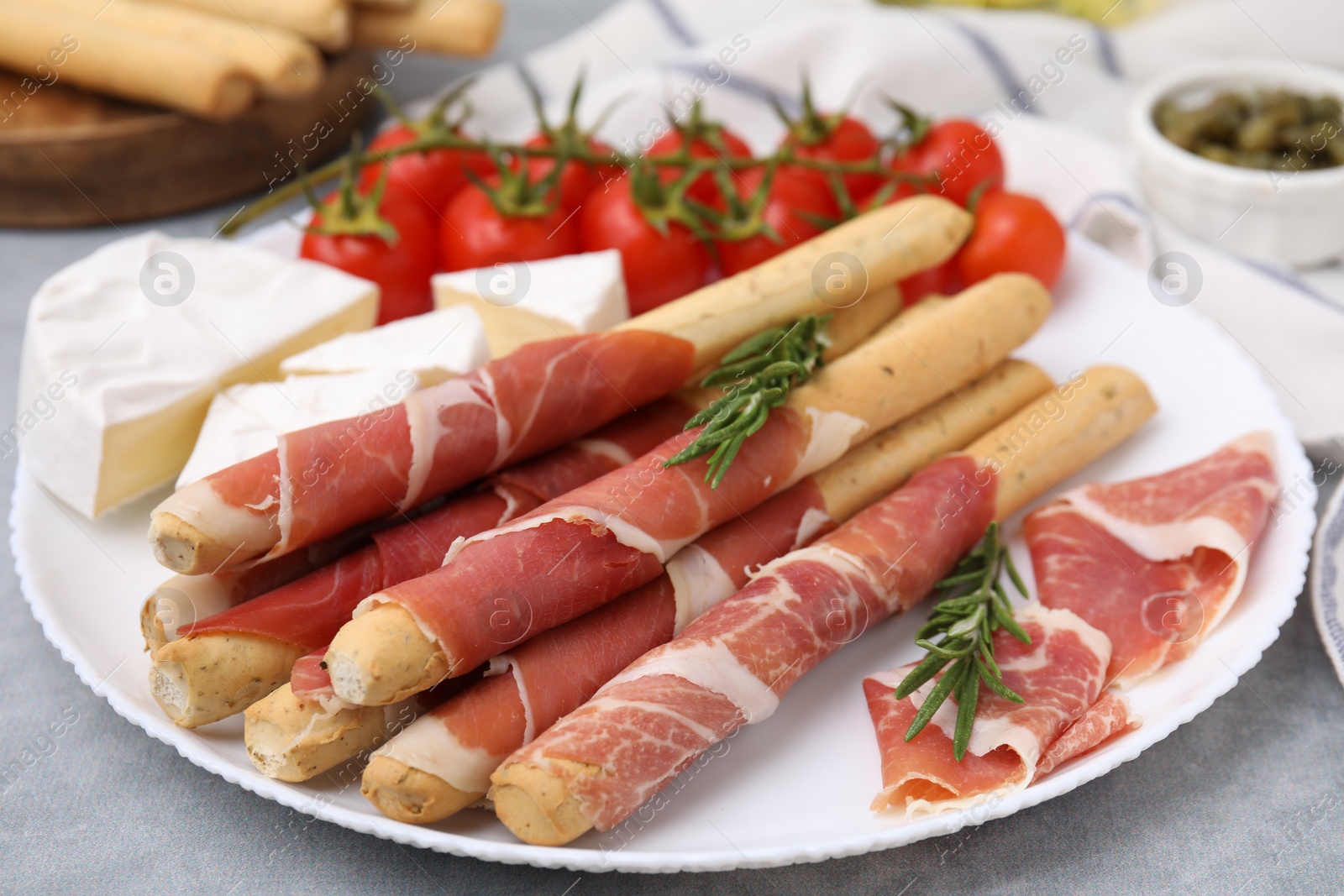 Photo of Plate of delicious grissini sticks with prosciutto, cheese and tomatoes on light grey table, closeup