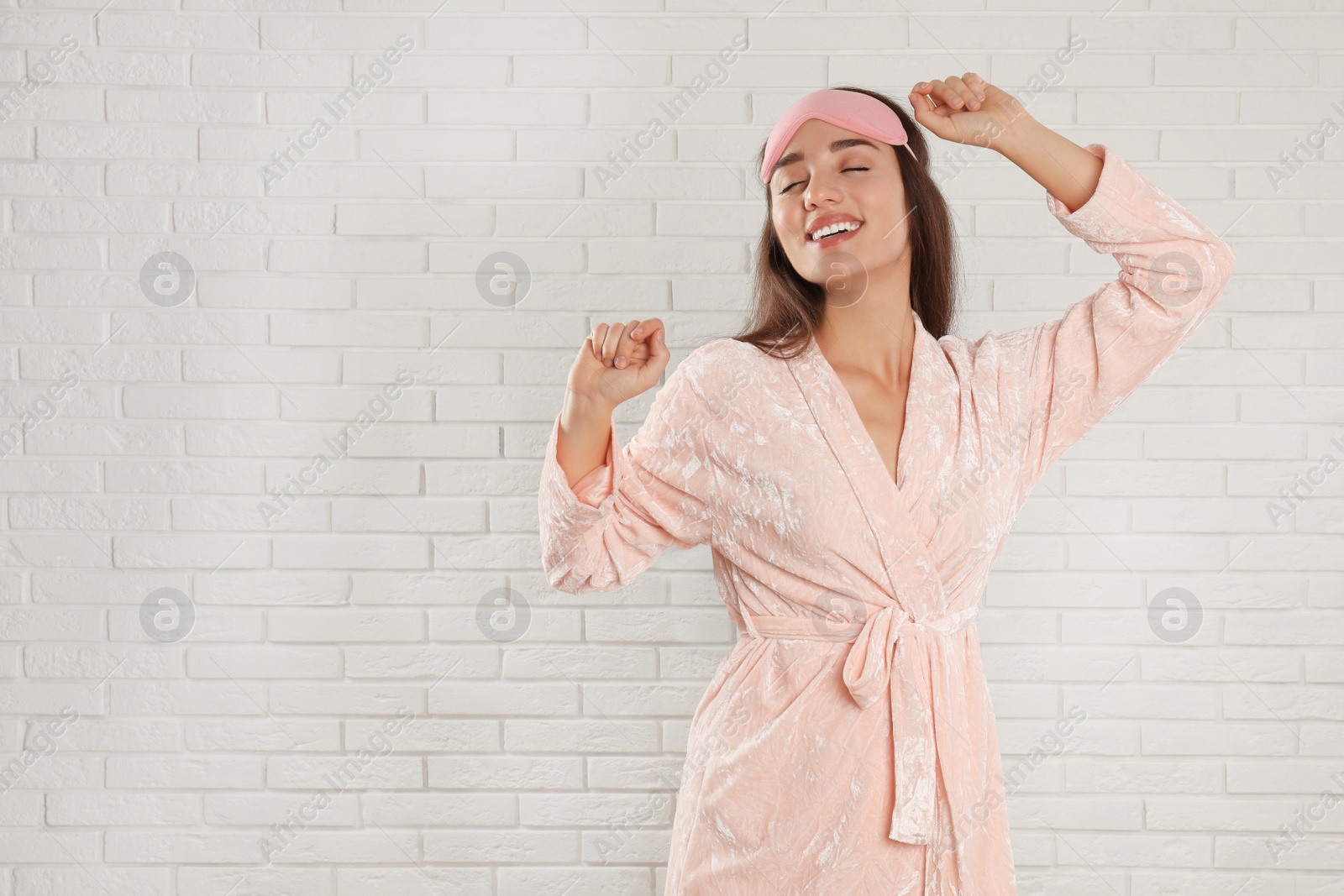 Photo of Young woman in bathrobe and eye sleeping mask near white brick wall. Space for text