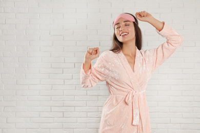 Photo of Young woman in bathrobe and eye sleeping mask near white brick wall. Space for text