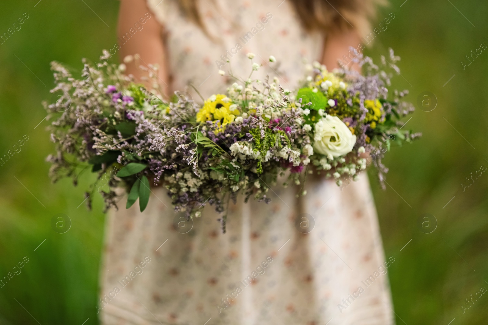 Photo of Cute little girl holding wreath made of beautiful flowers outdoors, closeup