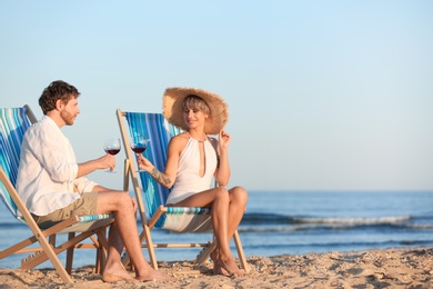 Photo of Young couple with glasses of wine in beach chairs at seacoast. Space for text