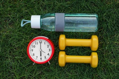 Photo of Alarm clock, bottle of water and dumbbells on green grass, flat lay. Morning exercise