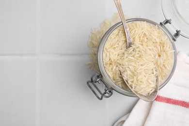 Photo of Glass jar and spoon with raw rice on white tiled table, top view. Space for text