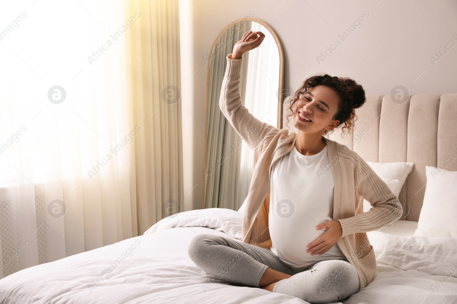 Image of Happy pregnant young African American woman sitting on bed in sunlit room