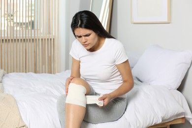 Young woman applying medical bandage onto knee in bedroom