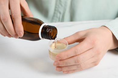 Photo of Woman pouring syrup from bottle into measuring cup at white table, closeup. Cold medicine