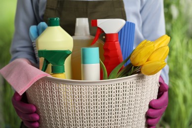 Photo of Woman holding basket with spring flowers and cleaning supplies outdoors, closeup