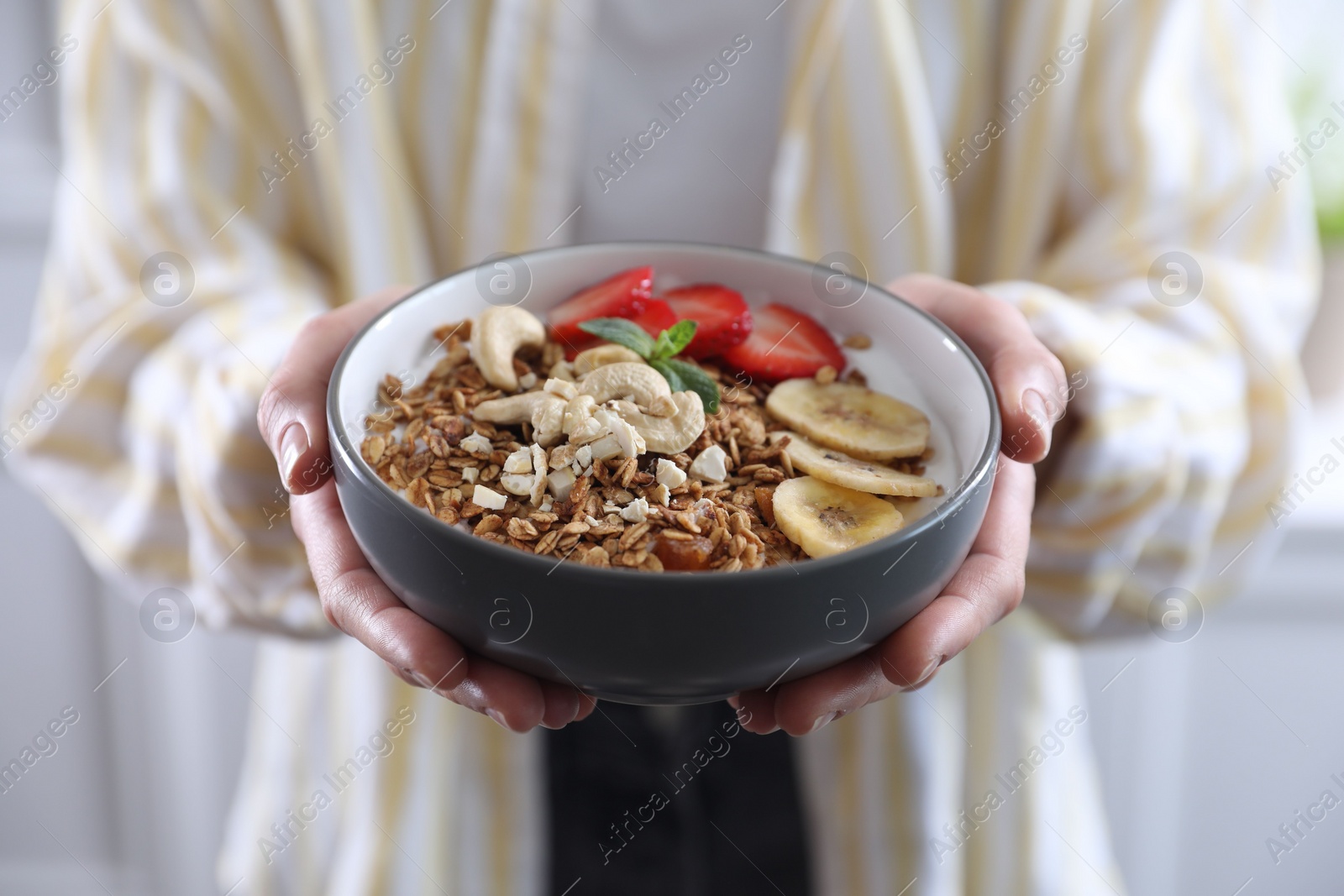 Photo of Woman holding bowl of tasty granola with banana, cashew and strawberries indoors, closeup