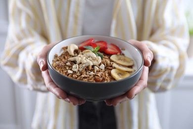 Woman holding bowl of tasty granola with banana, cashew and strawberries indoors, closeup