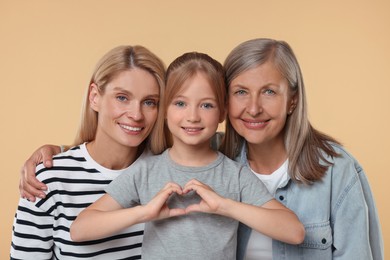 Photo of Three generations. Happy grandmother, her daughter and granddaughter making heart with hands on beige background