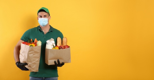 Photo of Courier in medical mask holding paper bags with food on yellow background, space for text. Delivery service during quarantine due to Covid-19 outbreak