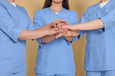 Photo of Nurses in medical uniforms putting hands together on light brown background, closeup