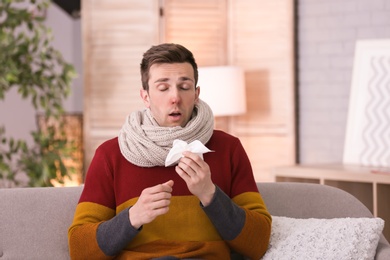 Sick young man with tissue suffering from cold on sofa at home