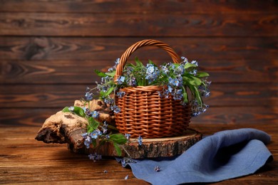 Beautiful forget-me-not flowers in wicker basket, piece of decorative wood and blue cloth on wooden table, space for text