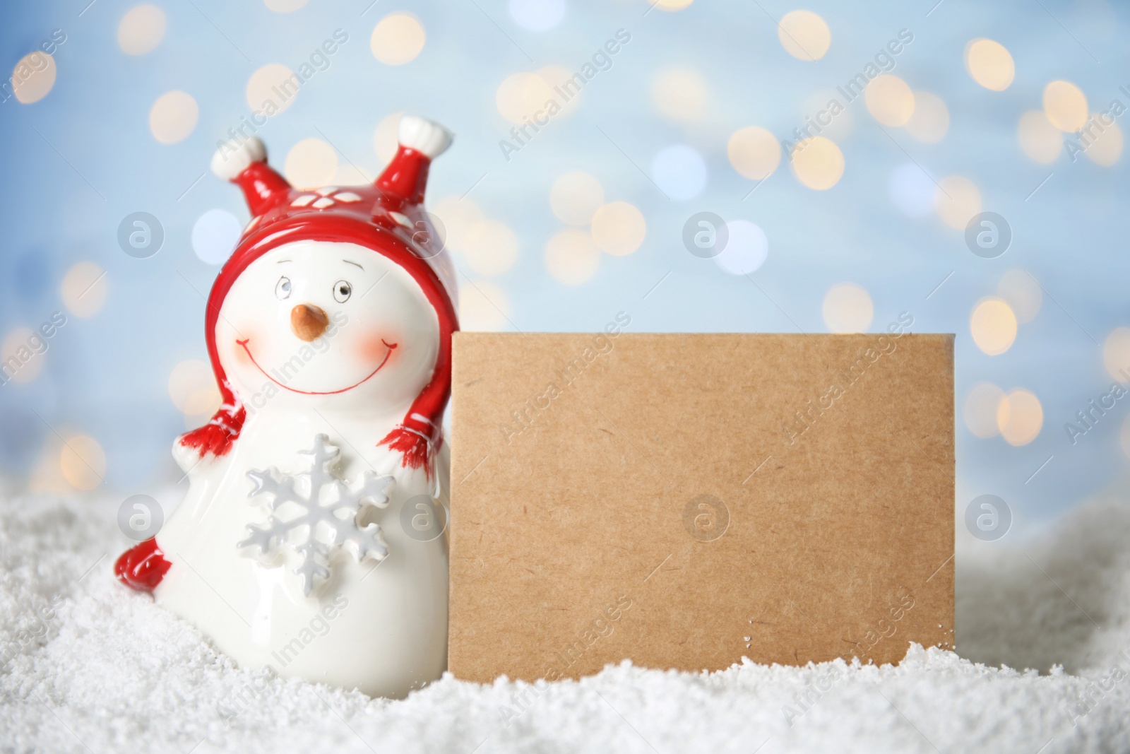 Photo of Decorative snowman near blank greeting card on artificial snow against blurred festive lights, space for text