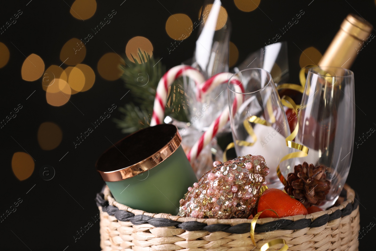 Photo of Wicker basket with Christmas gift set on black background with festive lights, closeup