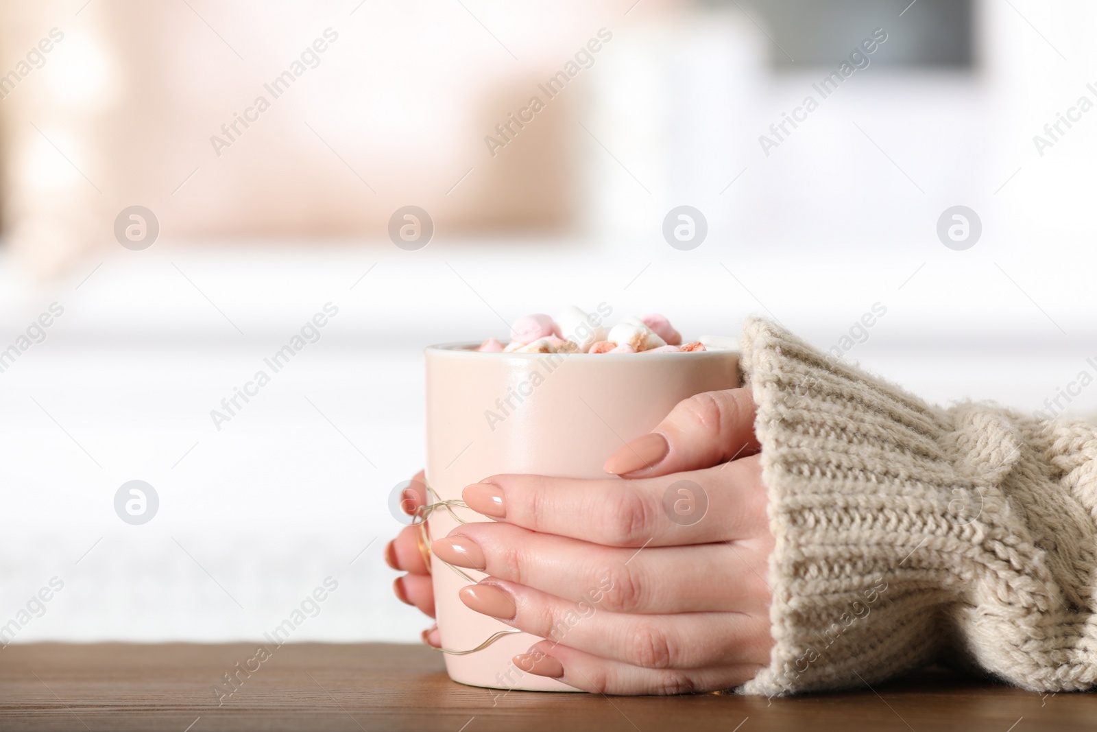 Photo of Woman in sweater holding cup of aromatic cacao with marshmallows on wooden table, closeup. Space for text