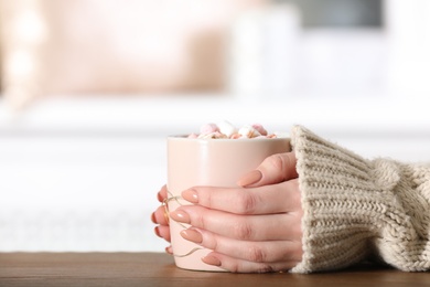 Photo of Woman in sweater holding cup of aromatic cacao with marshmallows on wooden table, closeup. Space for text