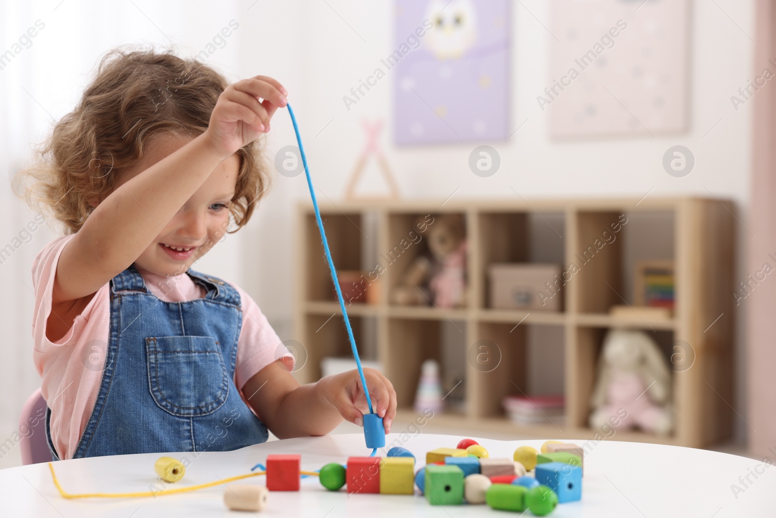 Photo of Motor skills development. Little girl playing with wooden pieces and string for threading activity at table indoors