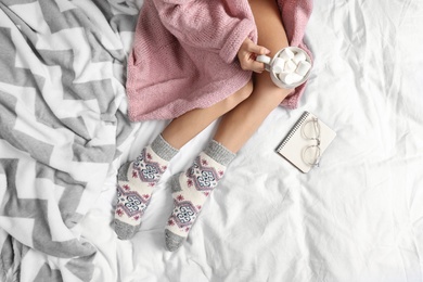 Photo of Woman with cocoa wearing knitted socks on white fabric, top view. warm clothes