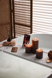 White wooden tray with smartphone, burning candles and beauty products on bathtub in bathroom