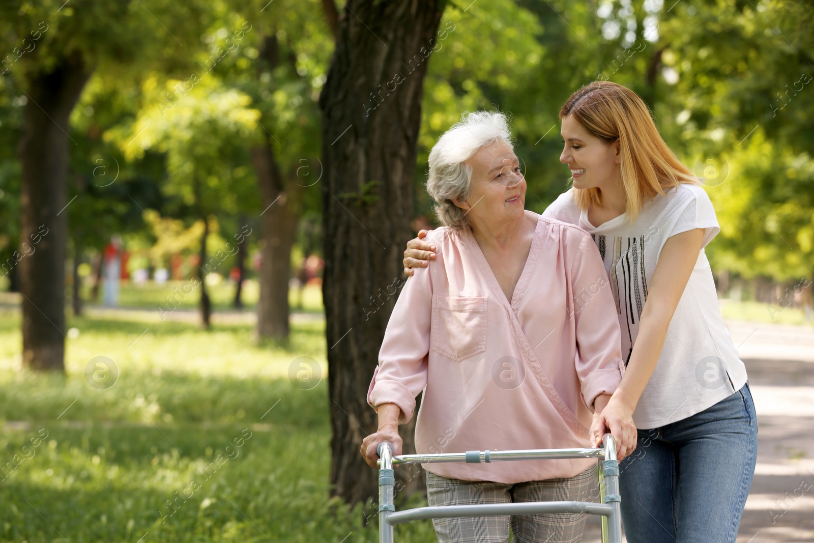 Photo of Caretaker helping elderly woman with walking frame outdoors