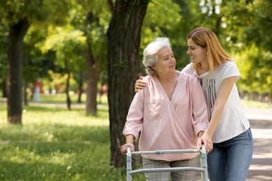 Photo of Caretaker helping elderly woman with walking frame outdoors