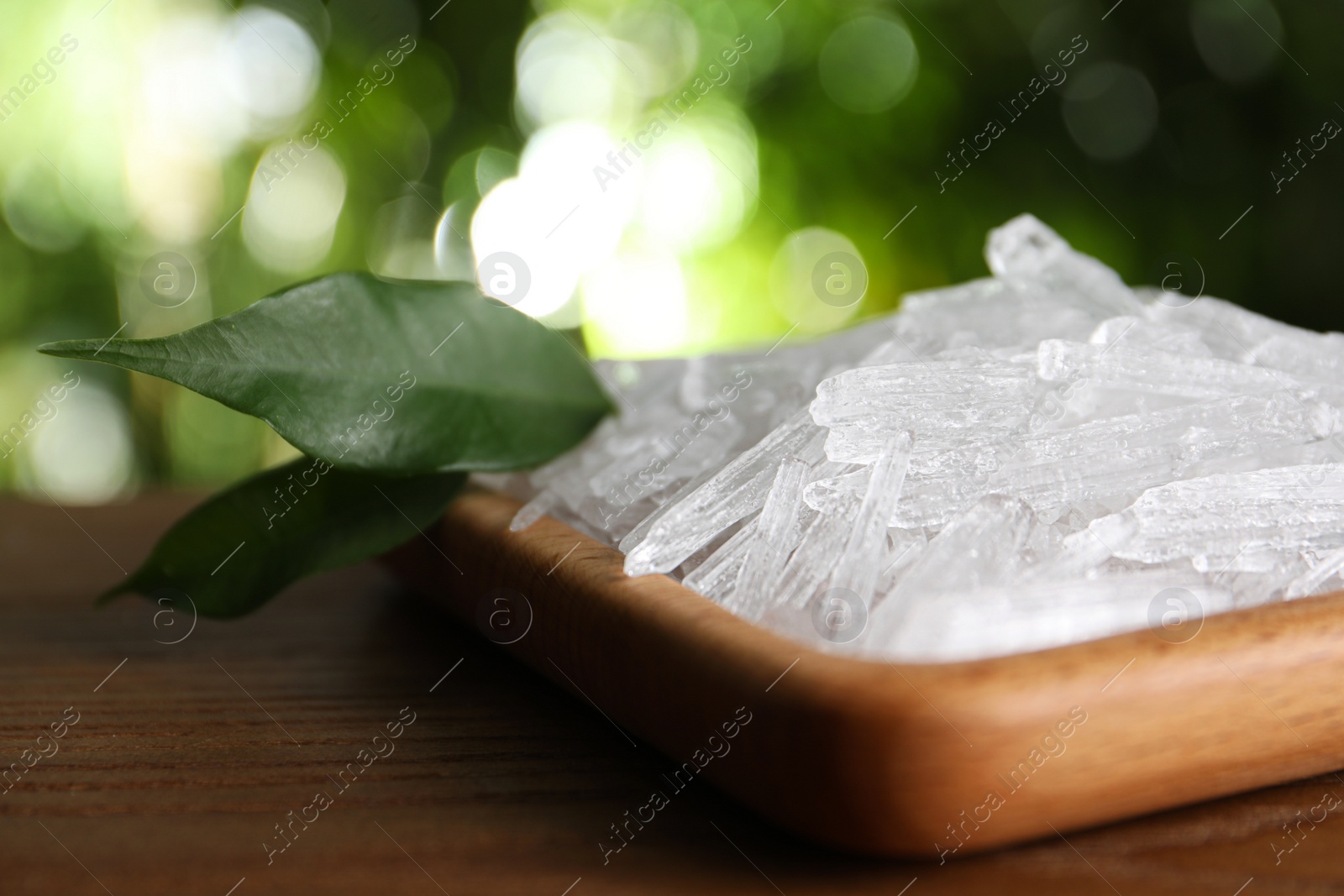 Photo of Menthol crystals and green leaves on wooden table against blurred background, closeup