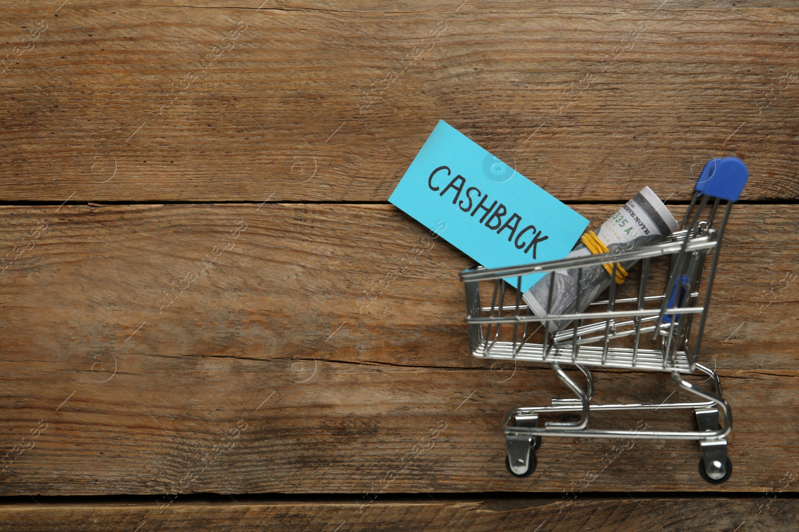 Photo of Card with word Cashback, rolled dollar banknotes and shopping cart on wooden background, flat lay. Space for text